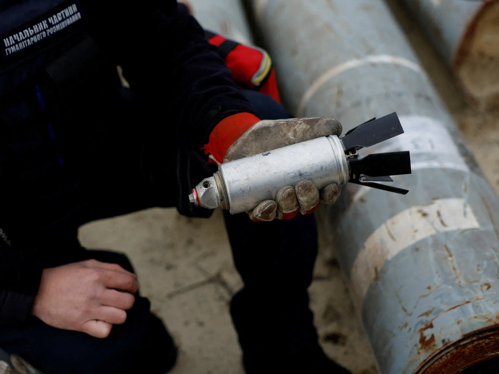 Gloved person holds a cylindrical munition beside a large industrial pipe.