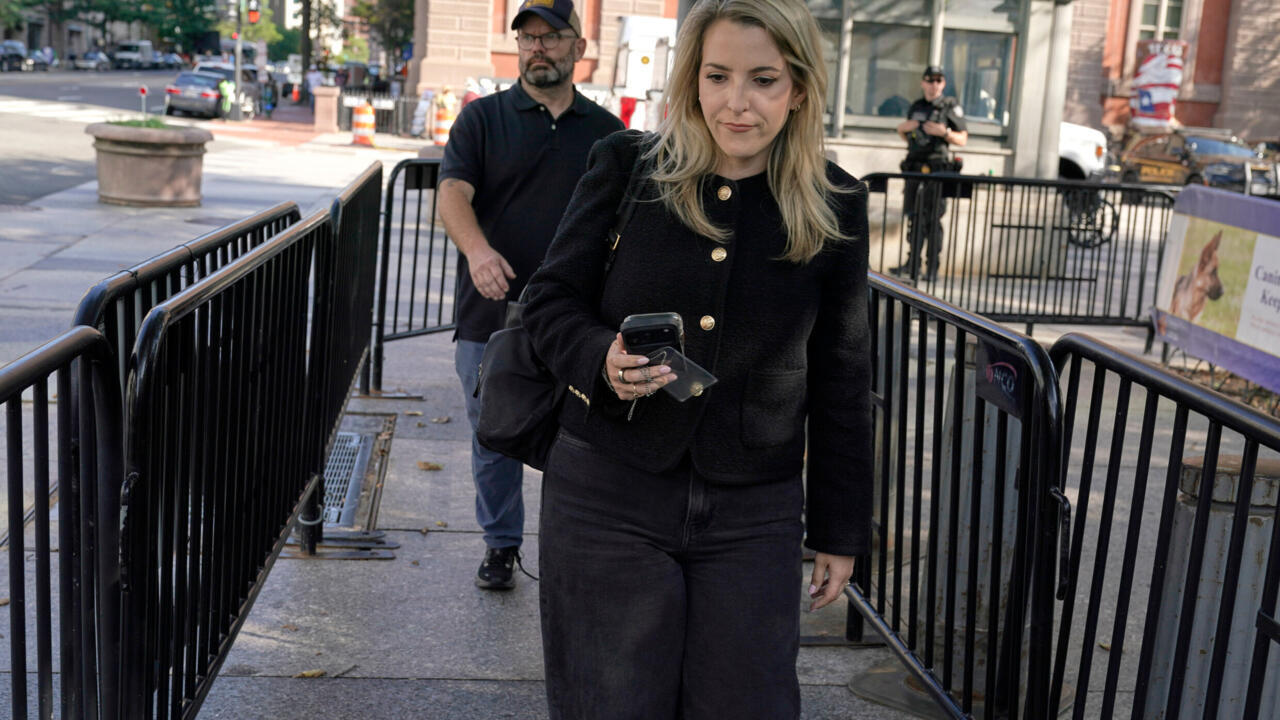 Blonde woman holding a phone walks between metal barricades as a man follows behind.