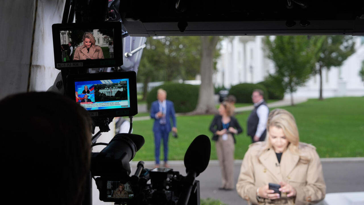 A camera crew films a blonde reporter checking her phone outside a white building.