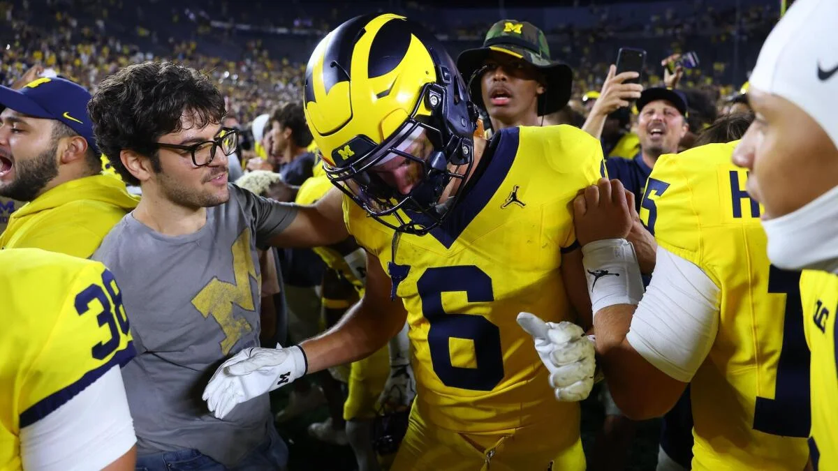 Michigan football player in yellow uniform and helmet celebrates amid teammates and fans on field