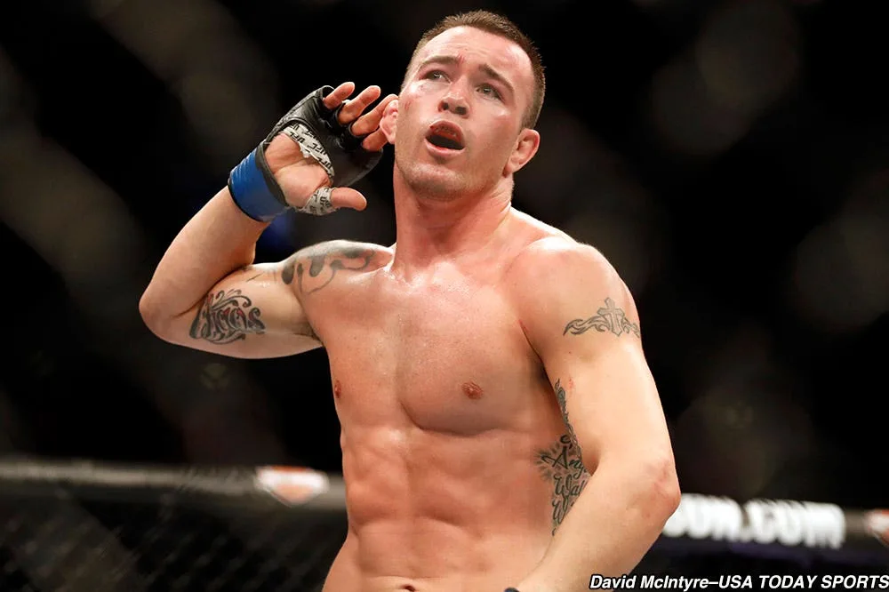 MMA fighter raises a gloved hand near his ear while standing inside a cage
