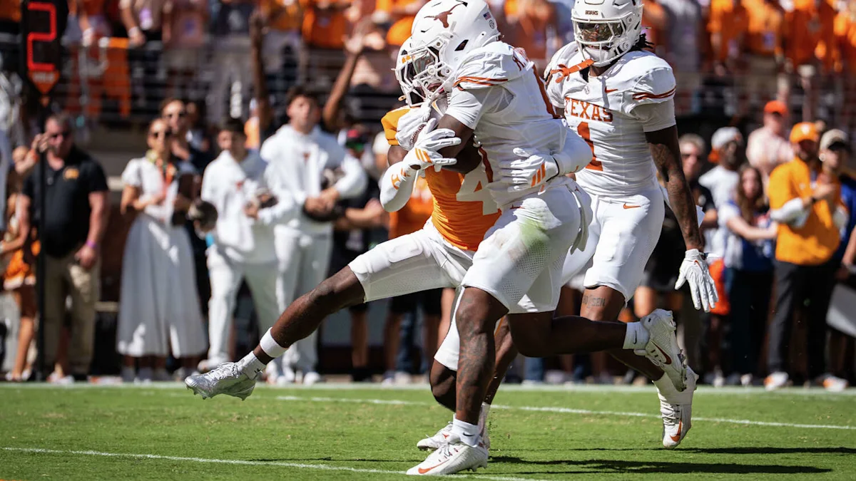 Texas players in white uniforms tackle an orange-clad ball carrier during a football game.
