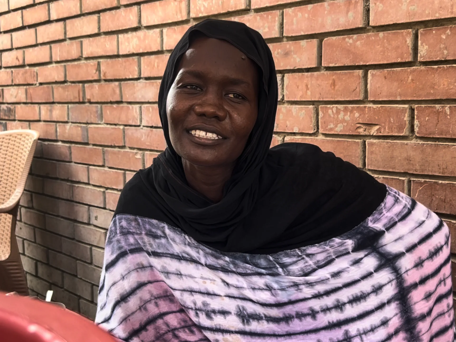 A smiling person in a black headscarf and patterned garment sits before a brick wall.