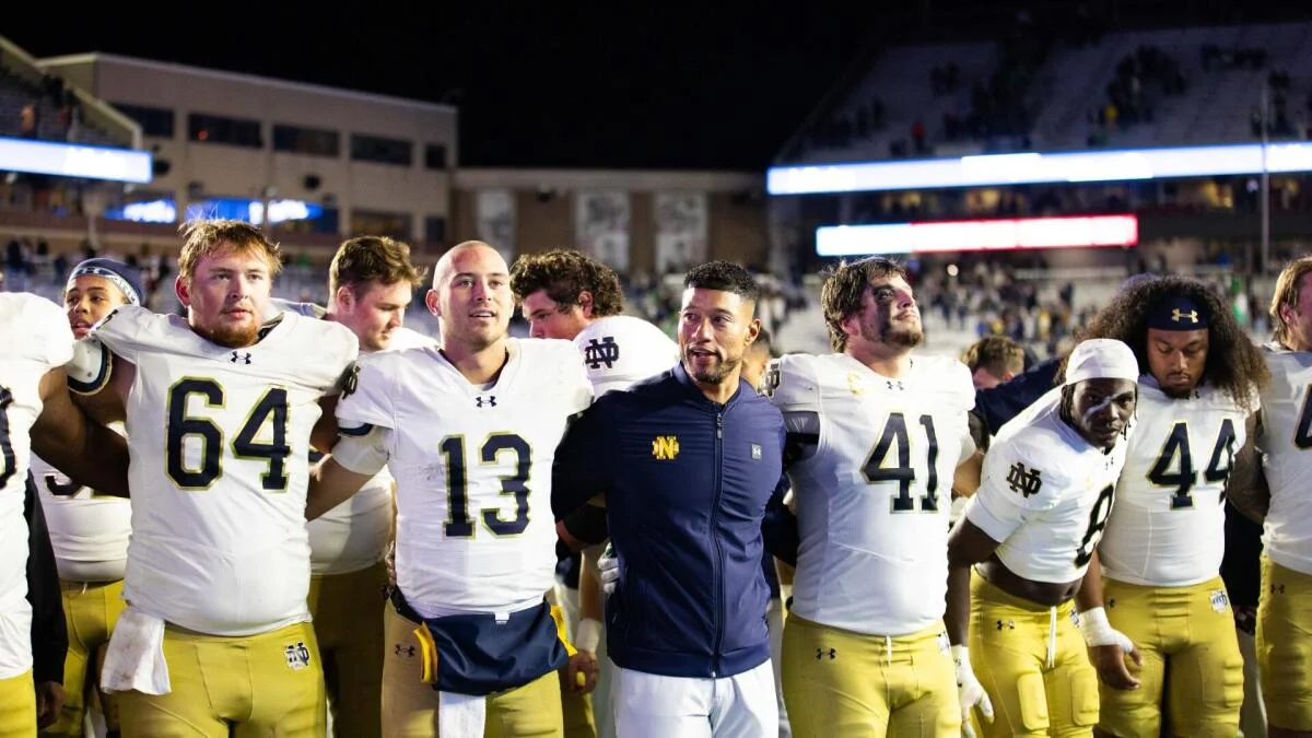 College football players and coach stand arm in arm on the field during a nighttime game.