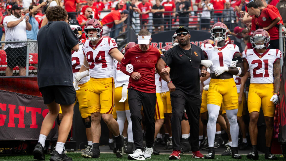USC football players and coaches walk onto the field surrounded by a crowd in red.