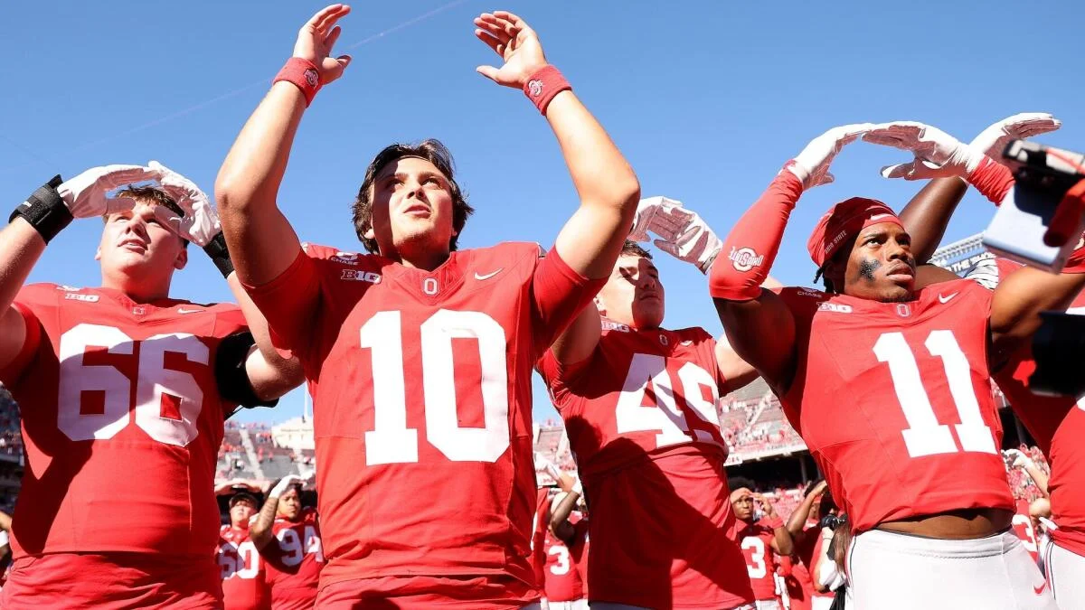 Ohio State players in red uniforms raise their arms beneath a clear blue sky.
