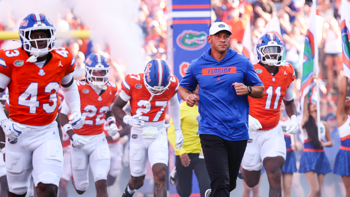 A coach in a blue Florida shirt jogs beside orange-uniformed football players entering the field.