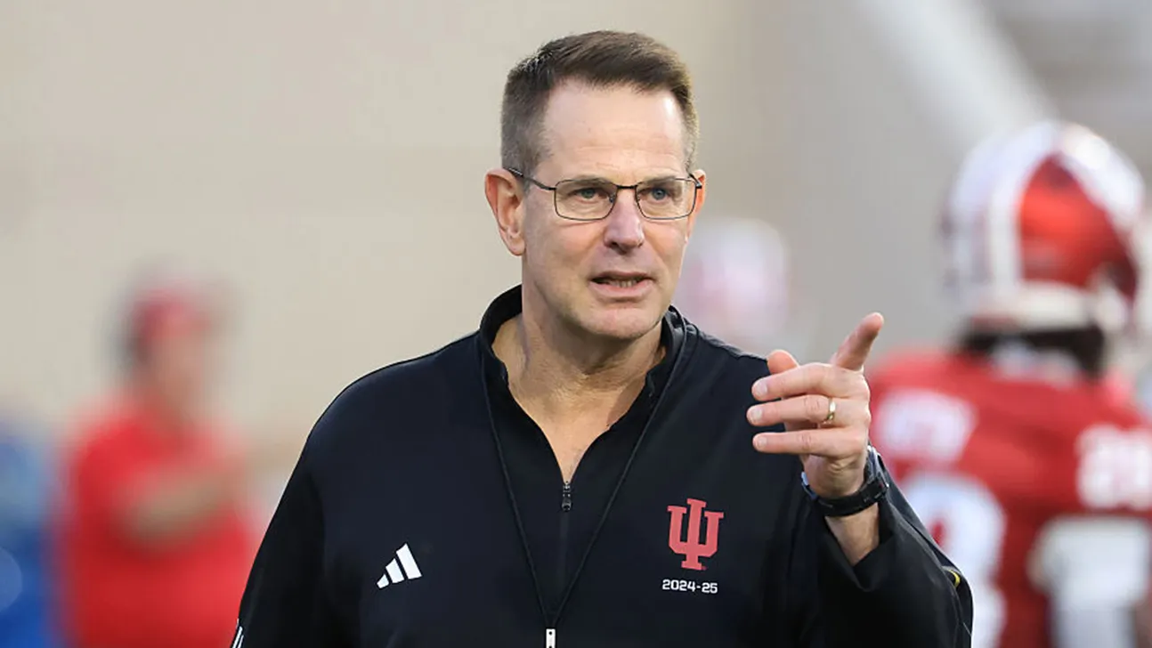 A football coach in an Indiana Hoosiers jacket points during a game, with players blurred behind him.