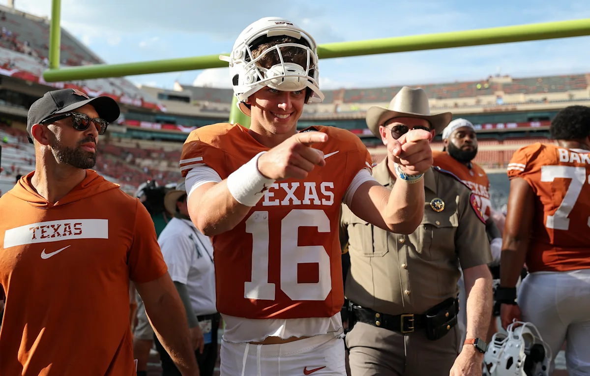 Football player in orange jersey and helmet points toward camera on field with teammates nearby