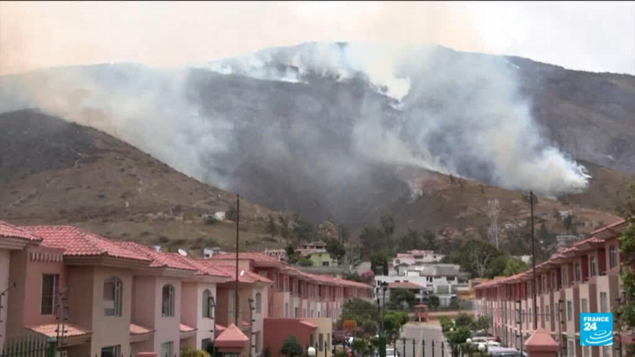 Smoke billows from a mountainside wildfire above rows of pink-roofed homes.
