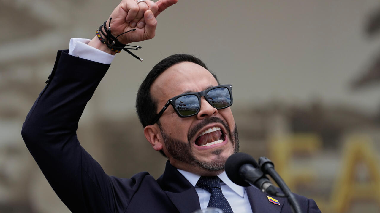Man in suit and sunglasses shouting into microphones with one fist raised