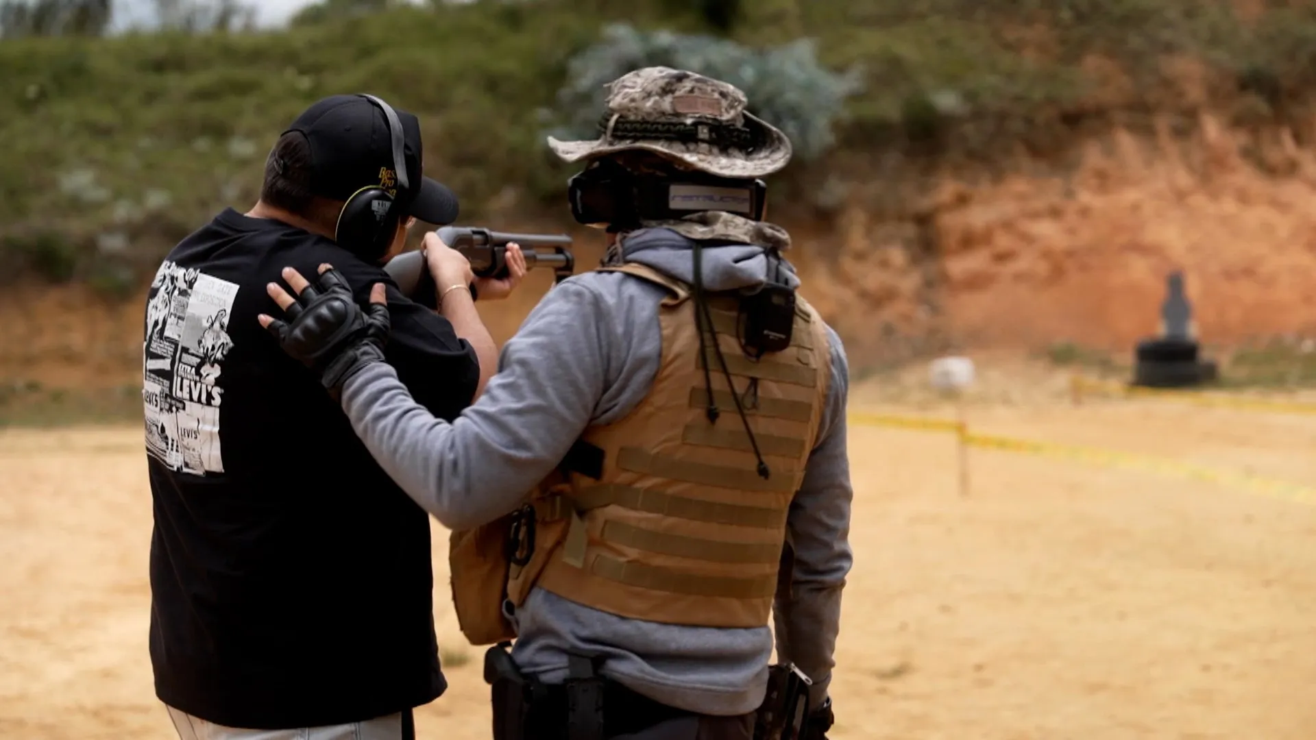 Two people stand at a shooting range, with one aiming a firearm under supervision.