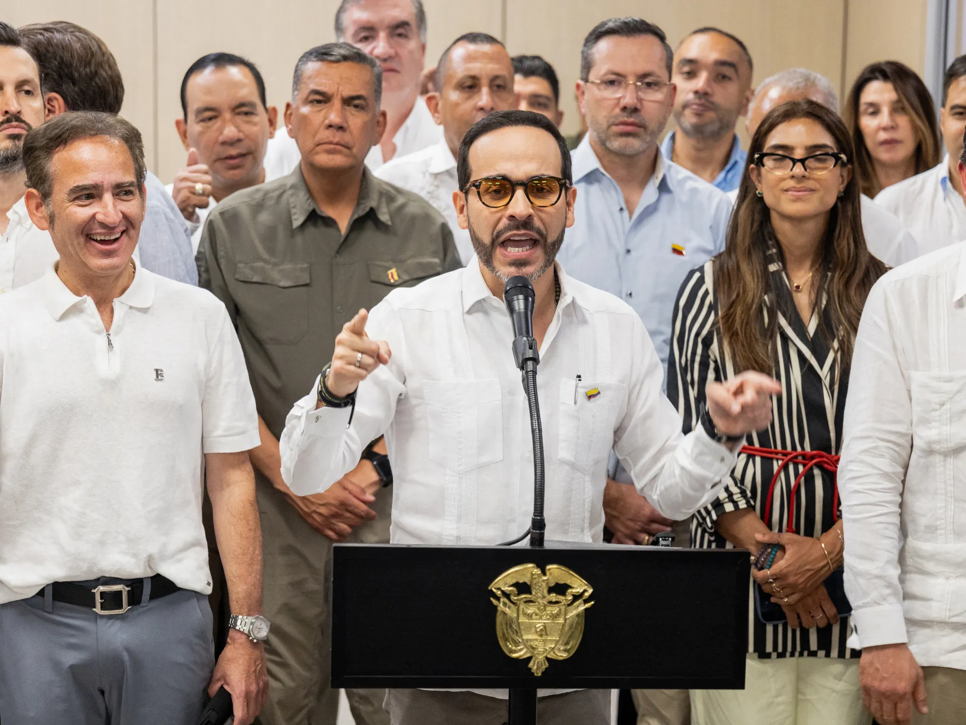 A man speaks at a microphone before a group of officials, with a national emblem on the podium.