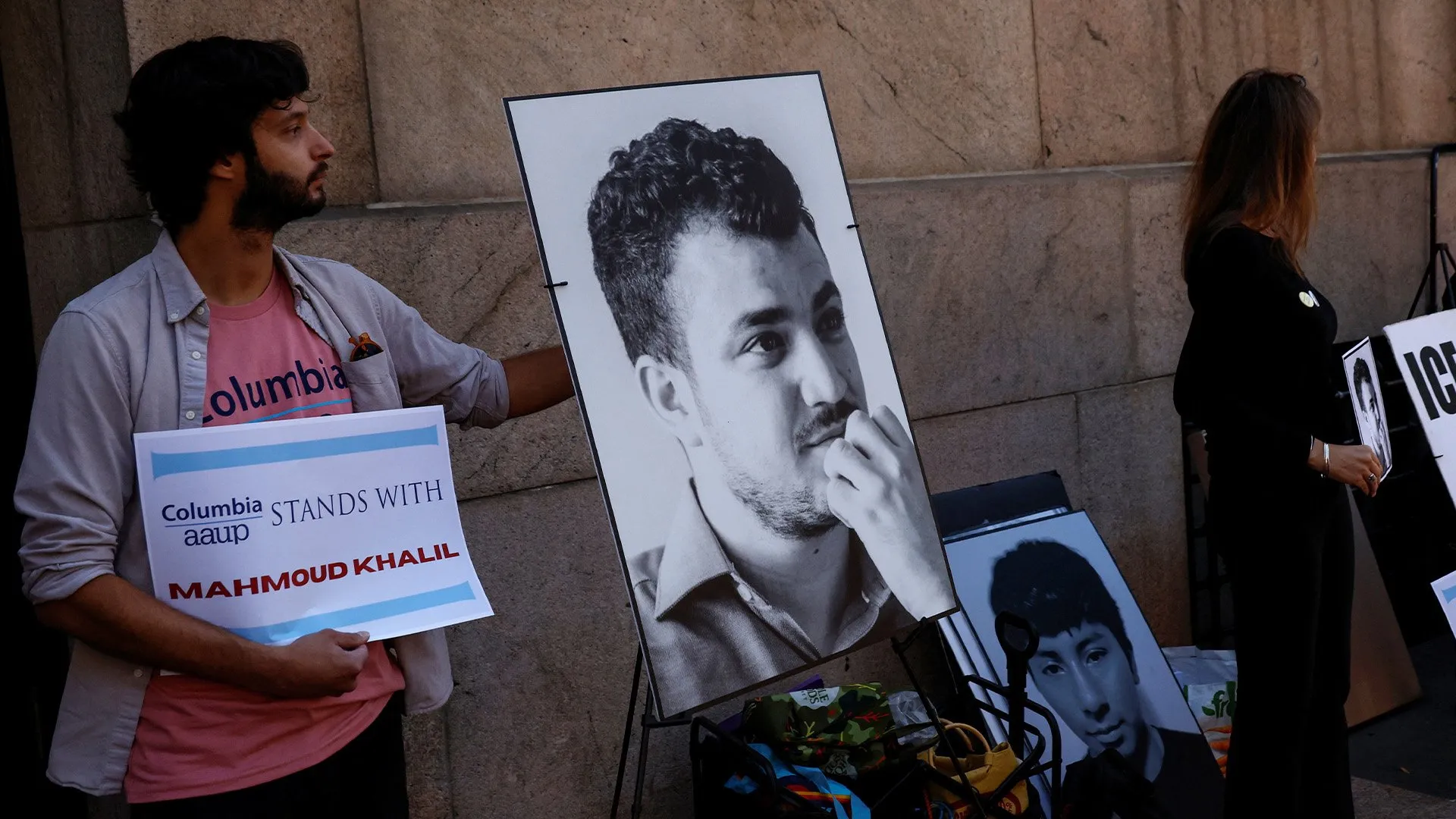 Protesters display portraits and signs supporting Mahmoud Khalil outside a stone building.