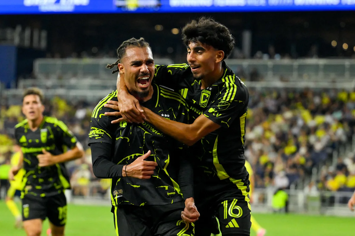Two soccer players in black and neon jerseys celebrate as a teammate approaches behind them.