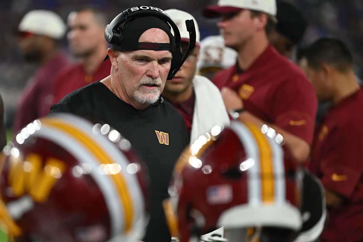 Football coach wearing a headset stands among players and helmets on the sideline.