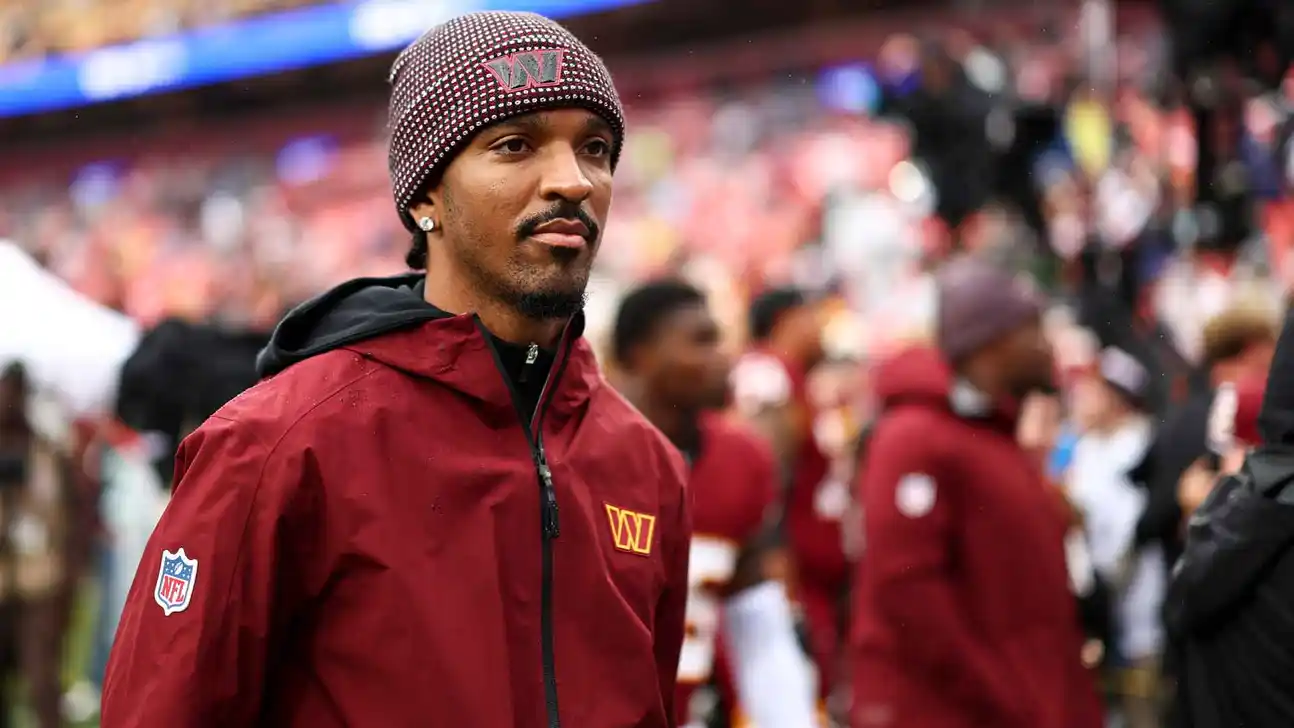 Man in a burgundy Washington Commanders jacket and knit beanie stands near a football field.