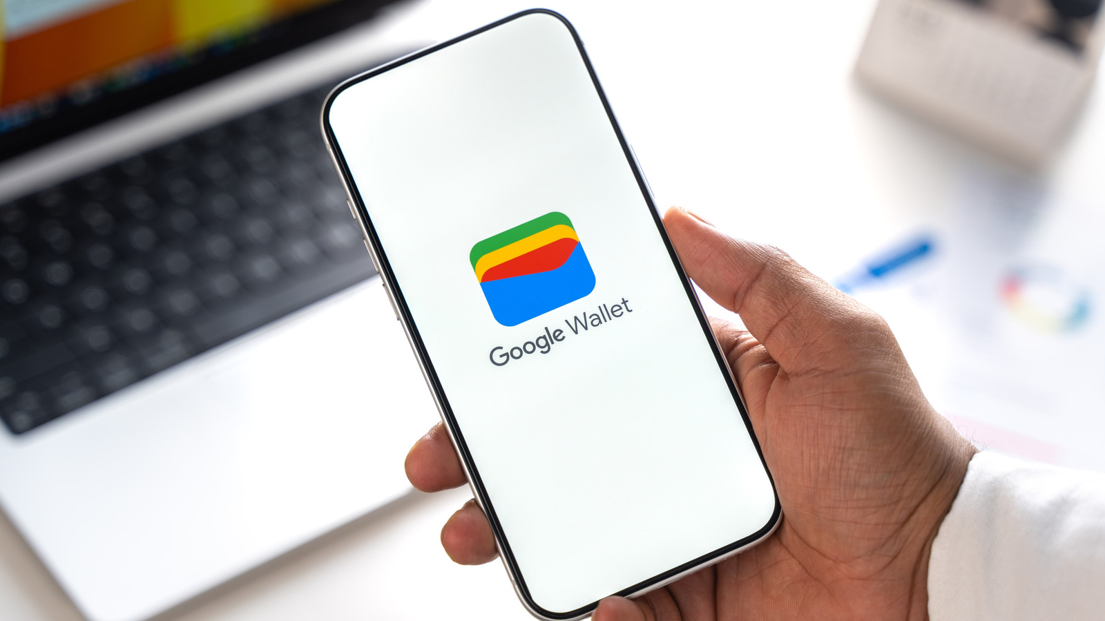 Hand holding smartphone displaying the Google Wallet logo above a blurred laptop keyboard