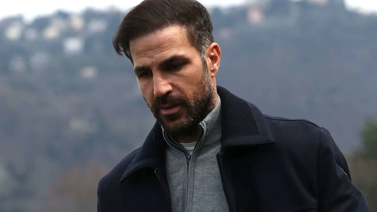 A bearded man in a dark coat and gray zip-up top stands outdoors against a blurred hillside.