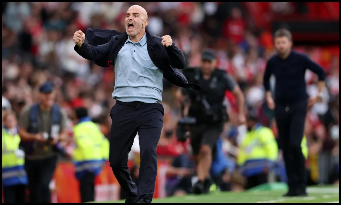 Bald football manager celebrates with clenched fists on the touchline during a match.
