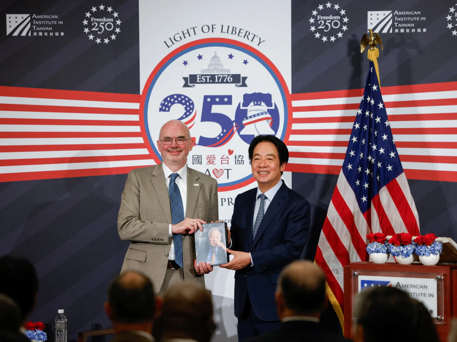 Two officials pose holding a booklet before a Taiwan-themed anniversary backdrop and American flags.