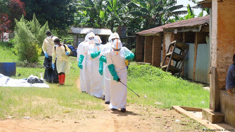 Health workers in protective suits walk along a rural path between brick buildings and banana trees.