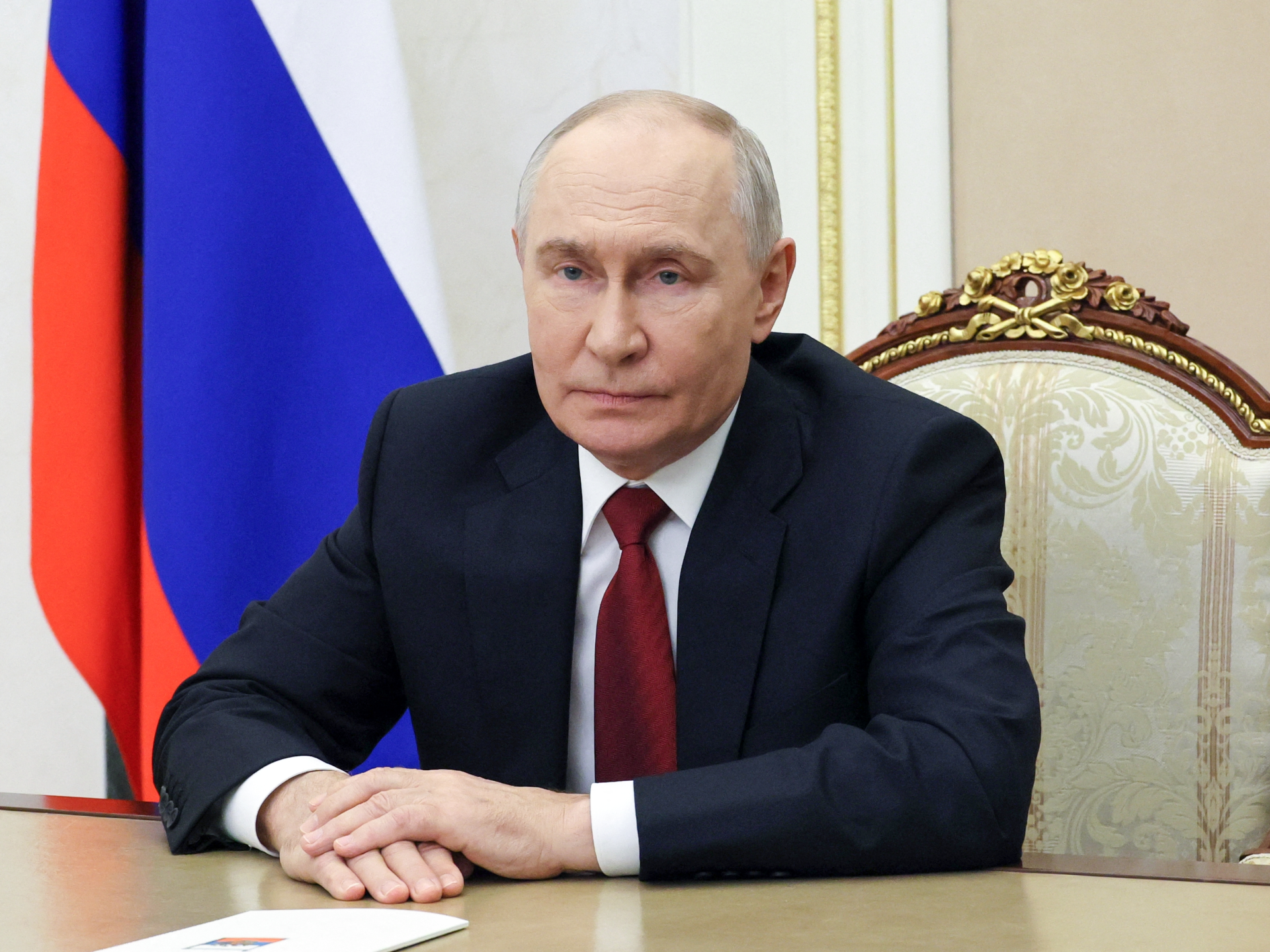 Man in dark suit seated at a desk with Russian flag and ornate chair behind him