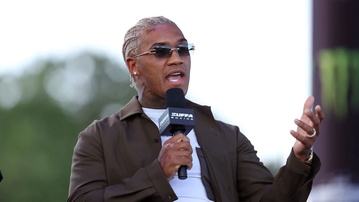 Man wearing sunglasses speaks into a Zuffa Boxing microphone while gesturing with his raised hand