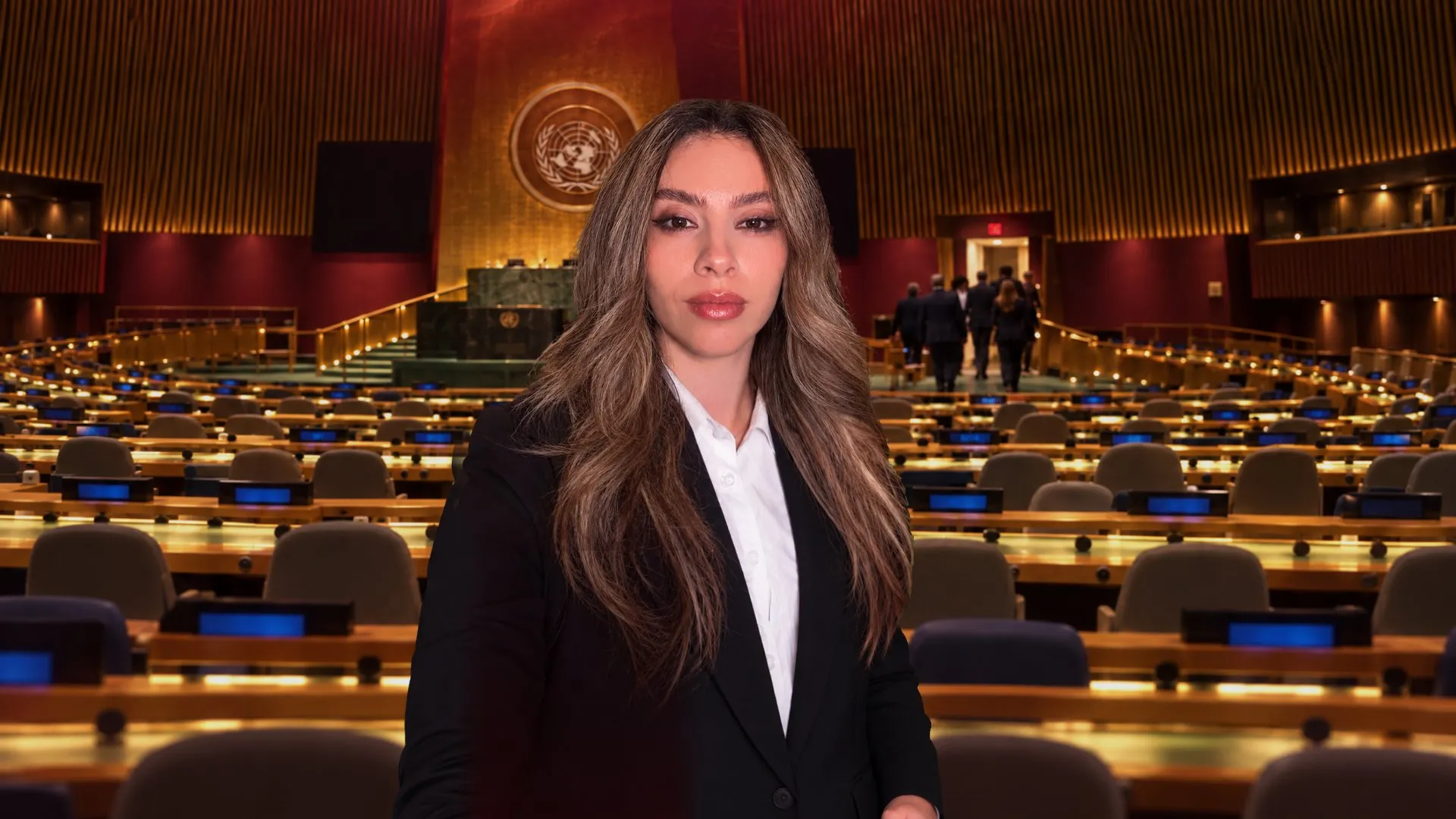 Woman in a dark suit stands before rows of desks in a large assembly chamber.