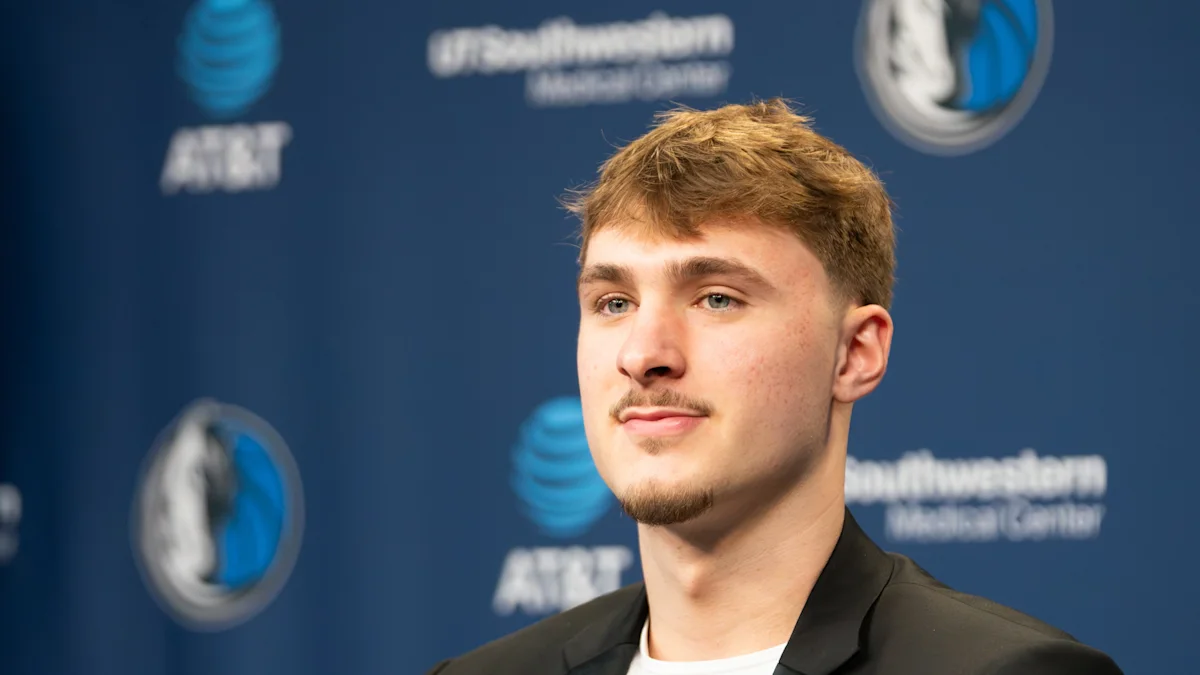 Young man in a black jacket poses before a blue backdrop with basketball team logos.