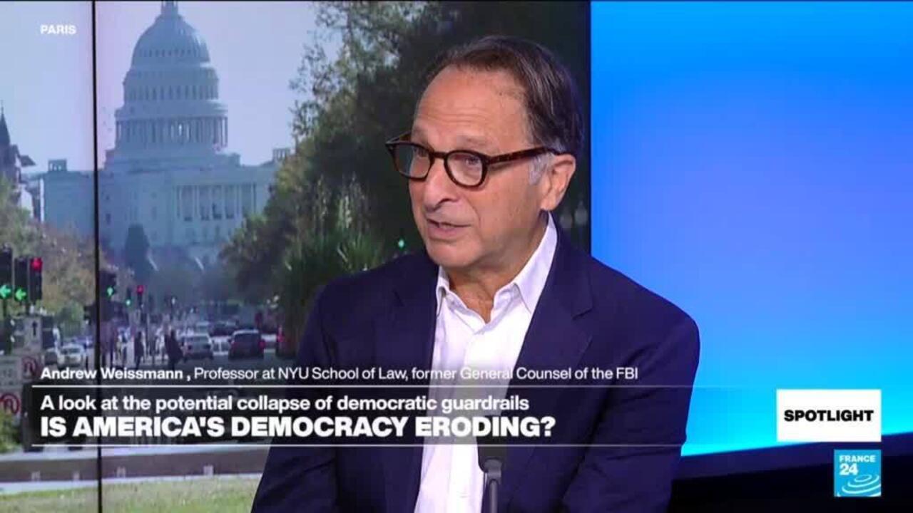 Man in glasses and suit speaks during a televised interview beside a capitol building backdrop.