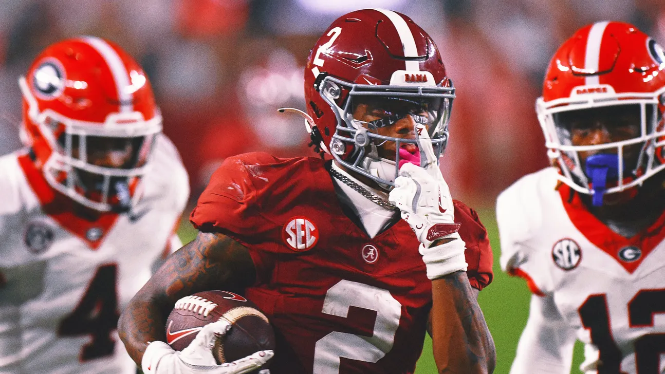 Alabama football player in red uniform carries a football between Georgia defenders.