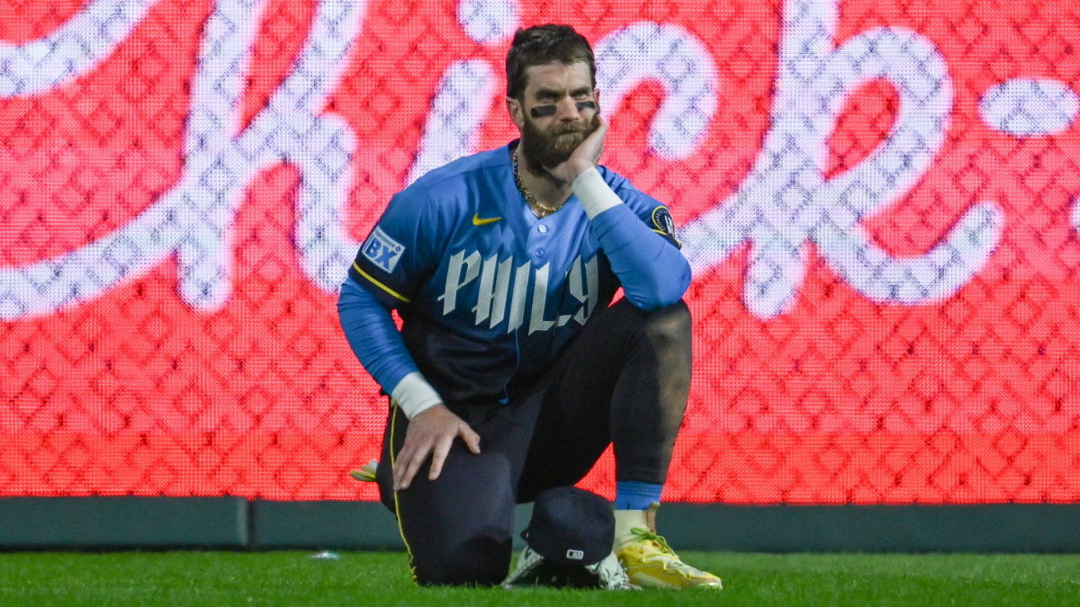 A bearded Phillies player kneels on the field beside his cap, with a bright red scoreboard behind him.
