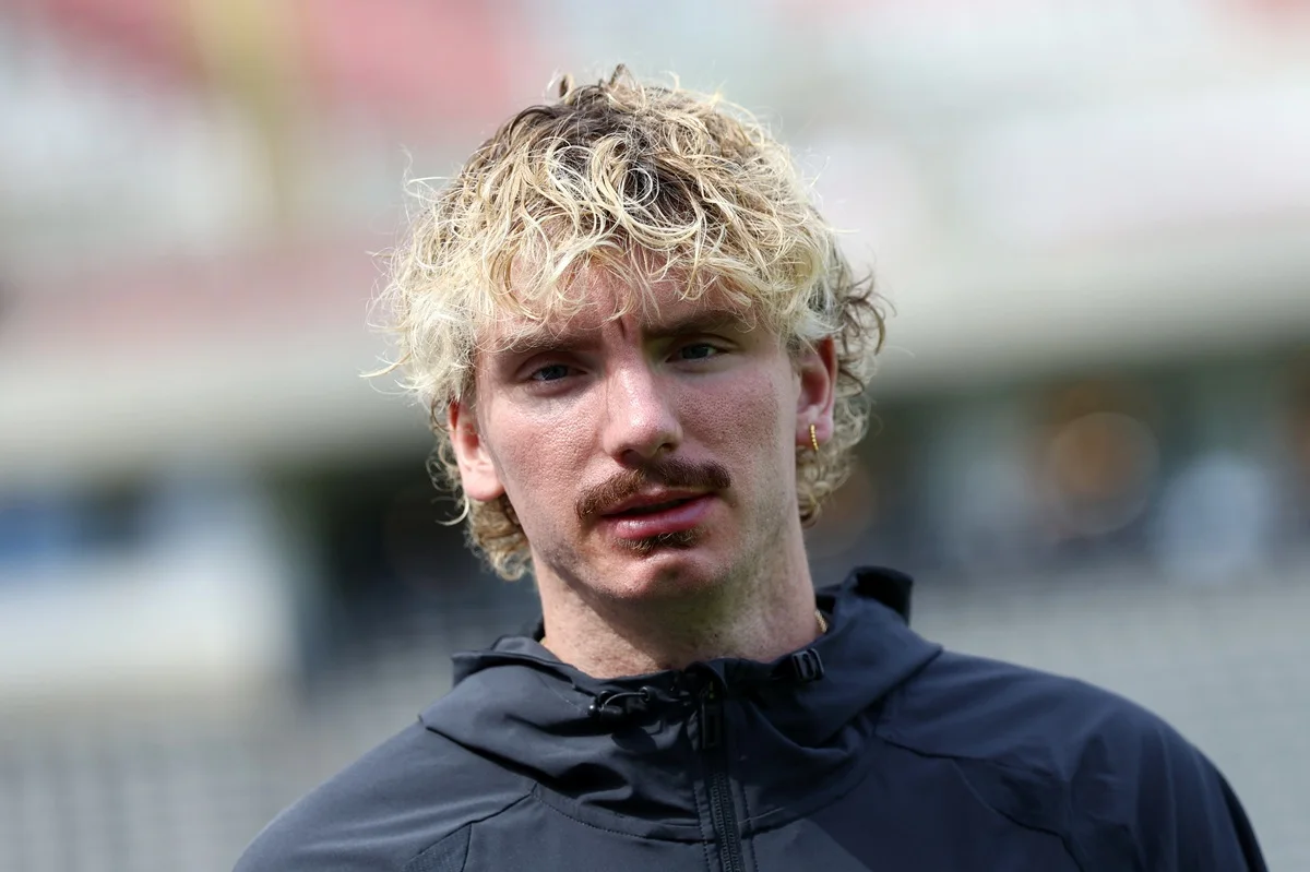 Blond-haired young man with mustache wearing a dark jacket outdoors