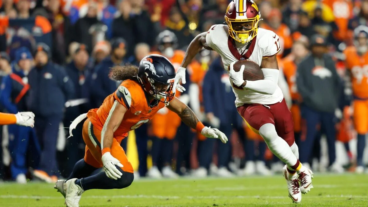 Football player in white and burgundy runs with the ball as an orange-clad defender reaches to tackle him
