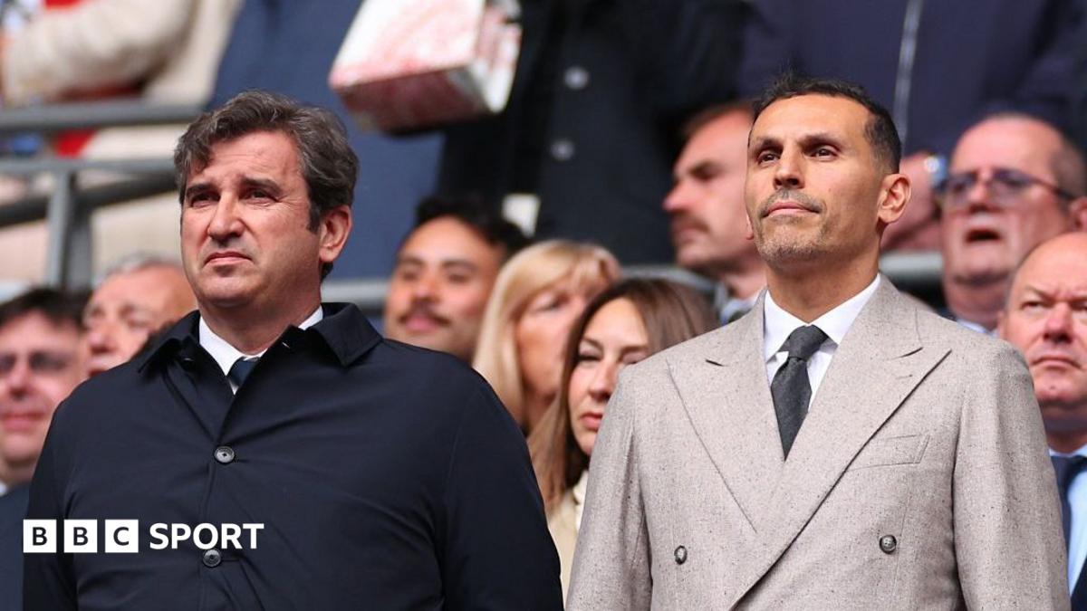 Two men in formal coats stand among spectators at a crowded sporting event.
