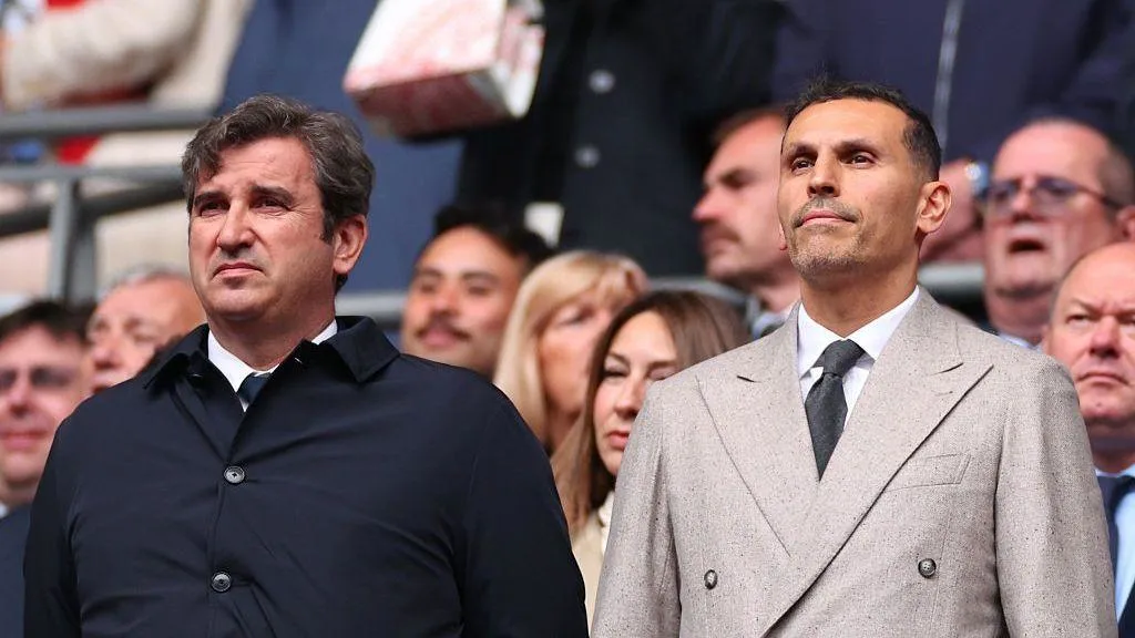 Two men in formal coats stand among spectators at a crowded sporting event.