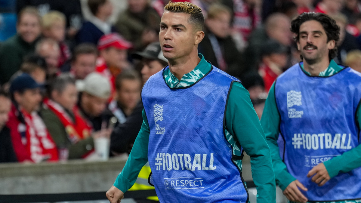 A footballer in a blue bib walks past spectators, with another bibbed player behind him.