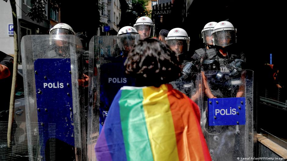 Helmeted police officers face a person draped in a rainbow-colored flag behind shields.