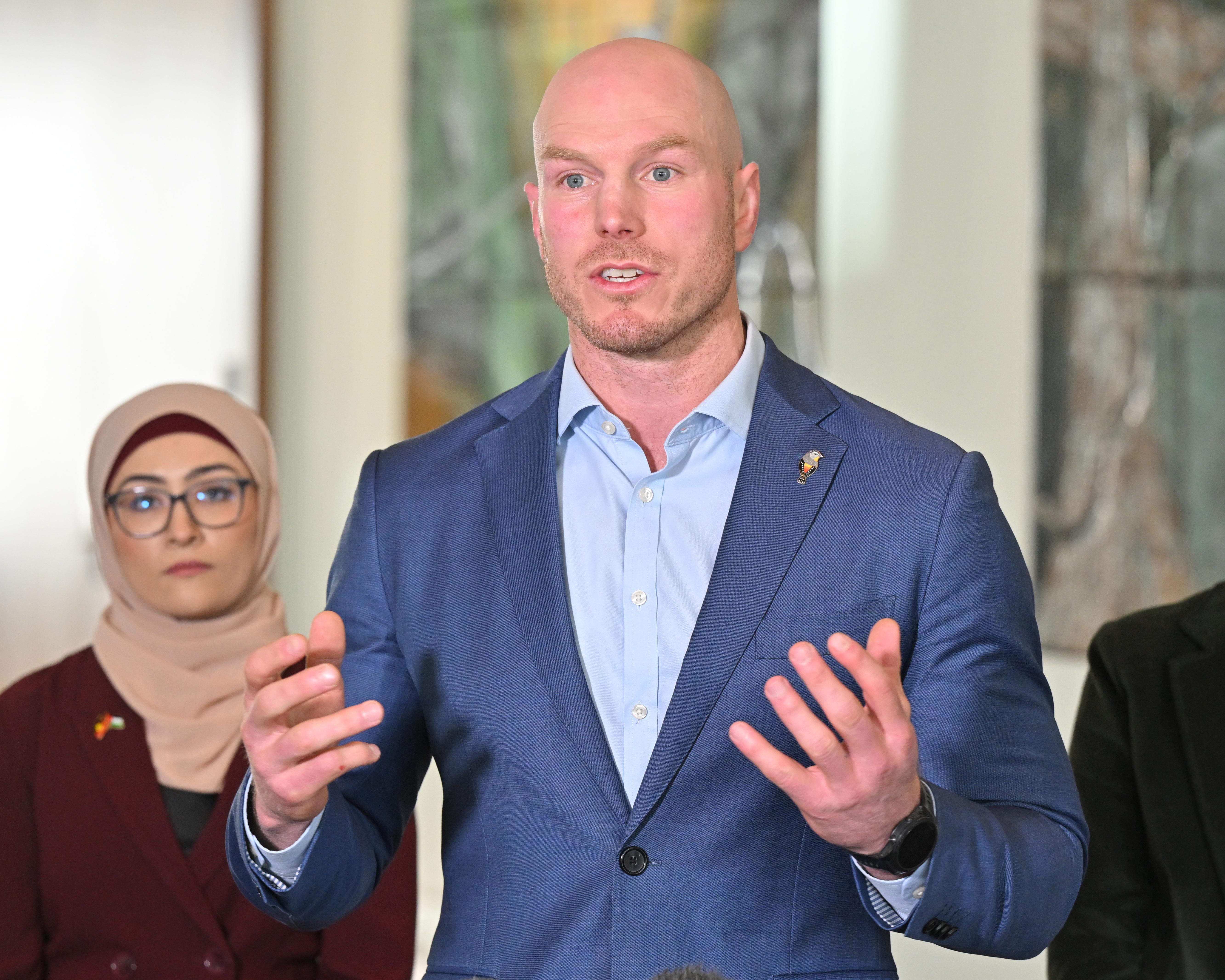 Bald man in blue suit gestures while speaking, with two people standing behind him indoors.