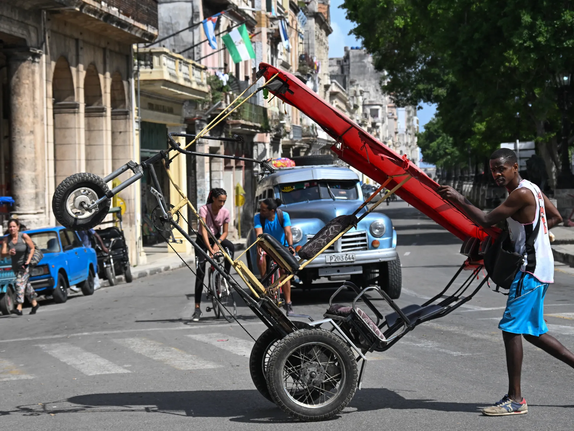 Man pushes a large red wheeled cart through a city street as vintage cars and pedestrians pass.