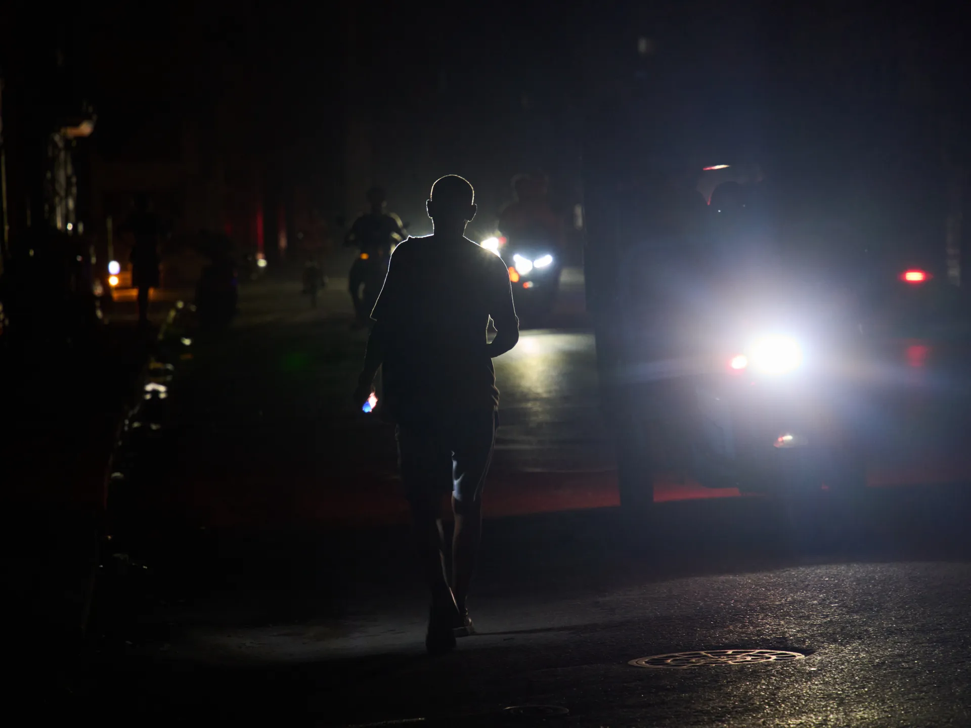 Silhouetted person walks along a dark street as approaching cars illuminate the nighttime scene.