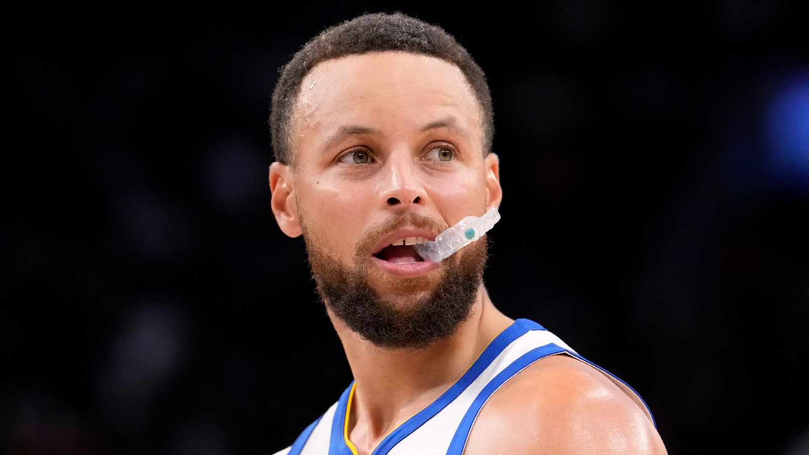 Basketball player in a white and blue jersey holds a clear mouthguard between his teeth during play.