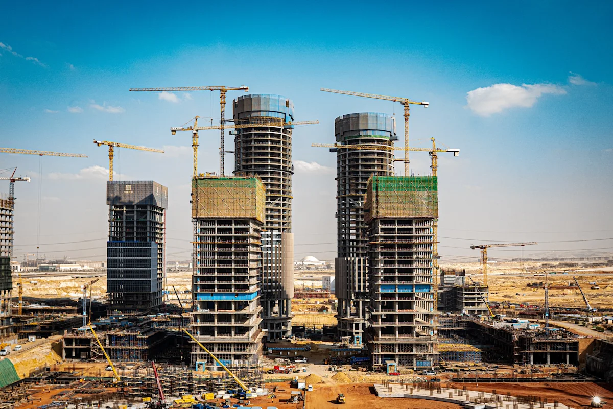 Multiple high-rise buildings under construction with cranes, scaffolding, and workers’ equipment beneath a blue sky.