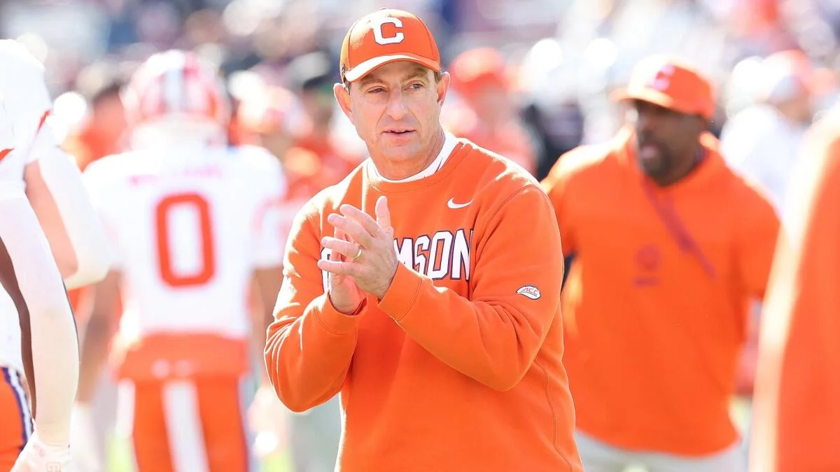 Clemson coach in orange cap and sweatshirt claps on the sideline during a football game.