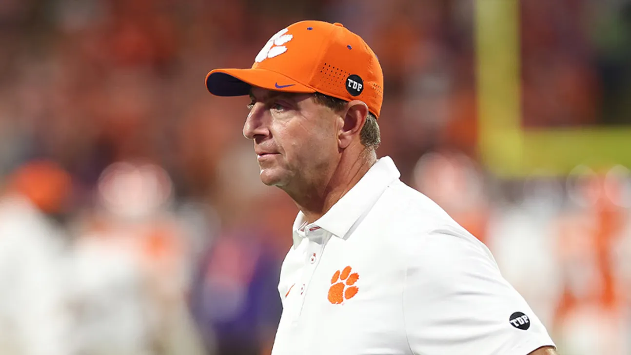 Clemson coach in an orange cap and white polo walks along the sideline.
