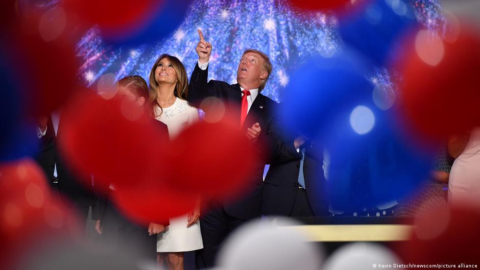Donald Trump points upward beside Melania Trump amid red and blue balloons and fireworks.