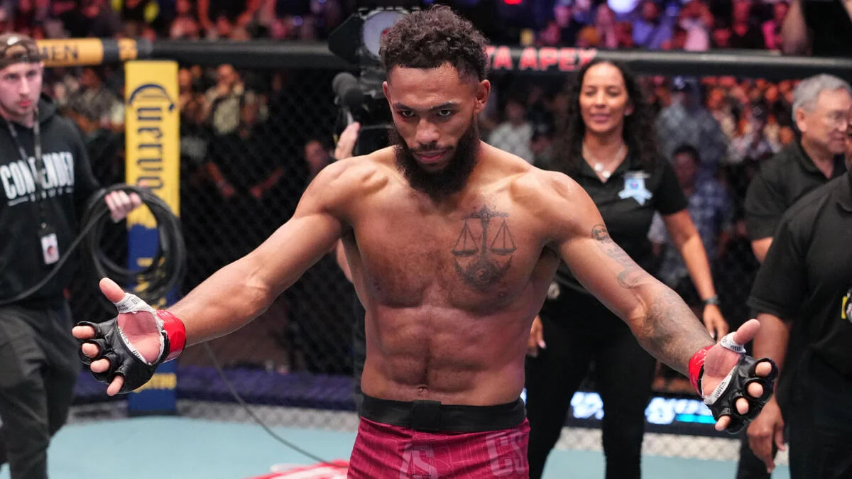 MMA fighter stands inside cage with arms outstretched, wearing red shorts and gloves