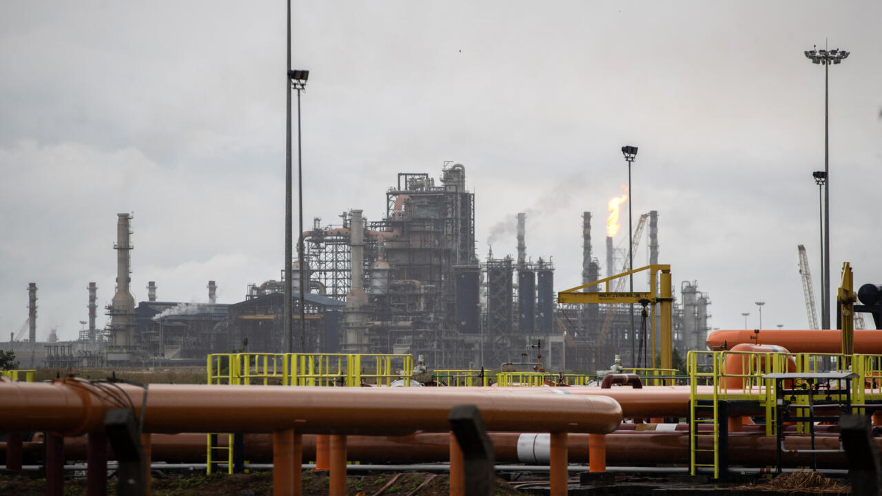 Industrial refinery complex with pipes, towers, cranes, and a bright flare beneath an overcast sky