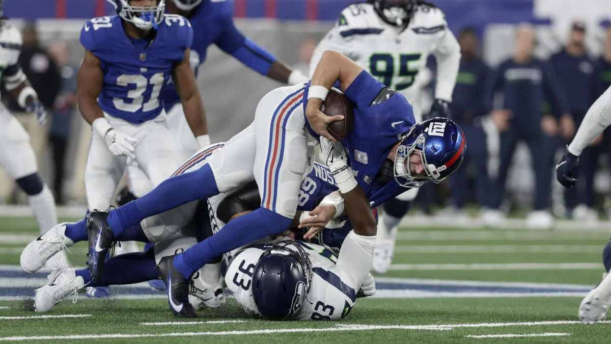 Quarterback in blue uniform is tackled by defenders in white uniforms on a football field